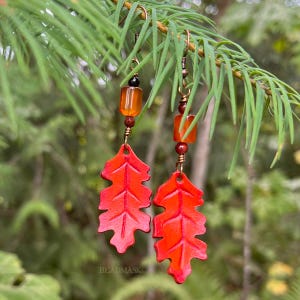 Peut inclure: Boucles d'oreilles artisanales avec des pendentifs en forme de feuilles rouges. Elles sont orn&eacute;es de perles orange et marron et de crochets cuivr&eacute;s. Le mot "BEADMASK" est visible en bas de l'image. Les boucles sont suspendues &agrave; une branche verte.