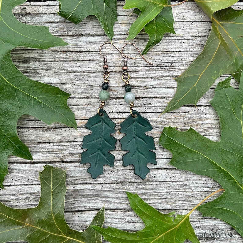 Peut inclure: Boucles d'oreilles en forme de feuilles vert fonc&eacute;, avec des crochets cuivr&eacute;s et des perles. Les boucles d'oreilles sont en mati&egrave;re textur&eacute;e, sur un fond en bois rustique avec des feuilles vertes.