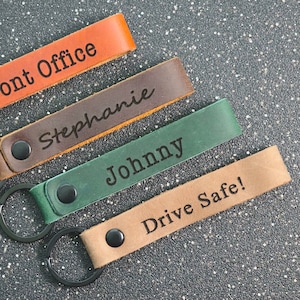Puede incluir: Cuatro llaveros de cuero con anillos de metal negro. Los llaveros son marrones, marrón oscuro, verdes y tostados. Los llaveros están grabados con el texto "Front Office", "Stephanie", "Johnny" y "Drive Safe!"