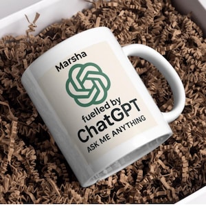 May include: White ceramic mug with the text "Marsha" and "fuelled by ChatGPT ASK ME ANYTHING" in black font. The mug features a green logo. The mug is inside a white gift box filled with brown paper shreds.