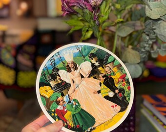 May include: A white ceramic plate with a colorful illustration of characters from the Wizard of Oz. The characters include Dorothy, the Scarecrow, the Tin Man, and the Cowardly Lion. The plate is decorated with a yellow brick road and a green field.