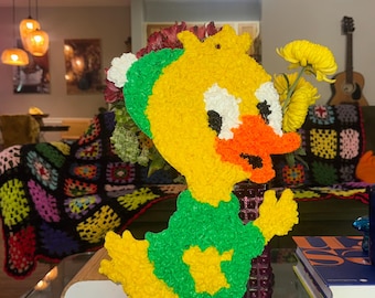 Op de afbeelding: Een getextureerde eendensculptuur in geel, groen en oranje. De eend heeft een groen vest, oranje voeten en snavel en witte accenten. Het is een decoratief item, mogelijk een vintage of handgemaakt stuk.