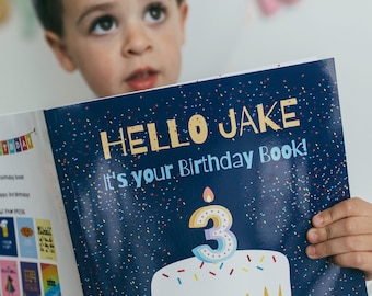 Pode incluir: Um livro de aniversário azul e branco com o texto "Hello Jake It's your Birthday Book!" na capa. O livro apresenta um fundo azul com confete colorido e um bolo branco com uma vela acesa em forma do número 3.
