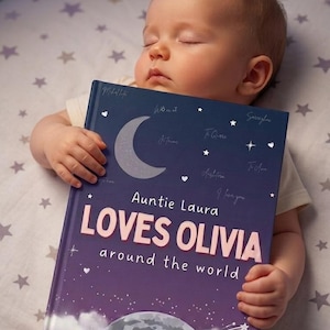 Puede incluir: Un bebé dormido sostiene un libro personalizado titulado "Auntie Laura Loves Olivia around the world". El libro tiene una cubierta azul oscuro con una luna creciente, estrellas y una ilustración del mundo.