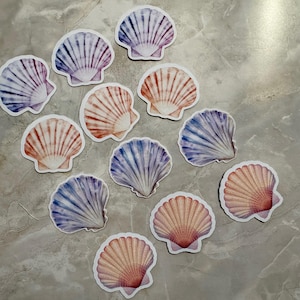 May include: A collection of seashell stickers in various colors, including shades of purple, orange, and blue. The stickers have a white border and are arranged on a gray surface. The stickers are a coastal or beach-themed craft supply.