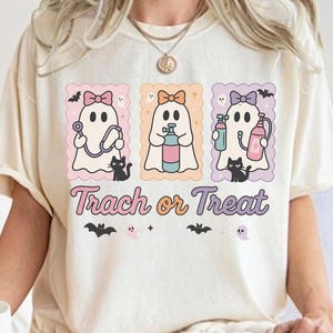 Puede incluir: Camiseta color crema con un diseño temático de Halloween. El diseño incluye tres fantasmas de dibujos animados con lazos, cada uno sosteniendo diferentes objetos médicos, acompañados de gatos negros y murciélagos. El texto "Trach or Treat" se muestra debajo.