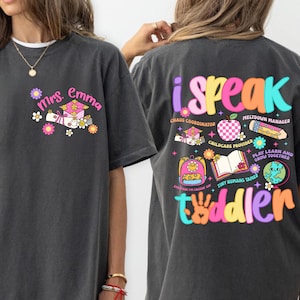 May include: A dark grey t-shirt with the text "Mrs. Emma" and a colourful graphic on the front. The back of the shirt features the words "i speak toddler" with additional text and graphics. The shirt is part of the Comfort Colors collection.