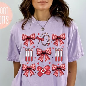 Puede incluir: Camiseta color orquídea con un gráfico que presenta lazos rojos, corazones, equipos médicos y vendajes. Las palabras "COMFORT COLORS" están en un círculo rosa en la parte superior izquierda. La palabra "ORCHID" está en la parte inferior derecha.