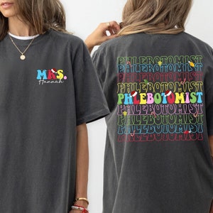 Puede incluir: Una camiseta gris con el texto "MRS. Hannah" en la parte delantera y el texto "PHLEBOTOMIST" repetido en colores arcoíris en la parte posterior.