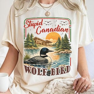 Pode incluir: Camiseta creme com a frase "Stupid Canadian WOLF BIRD". O design inclui uma cena de lago com um mergulhão, árvores e um pôr do sol. A camiseta tem um corte folgado.