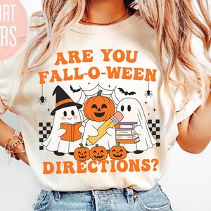 Puede incluir: Camiseta color crema con el texto "ARE YOU FALL-O-WEEN DIRECTIONS?" en naranja. El gráfico presenta fantasmas de dibujos animados, calabazas, un lápiz y libros, con telarañas y murciélagos. La camiseta tiene las palabras "COMFORT COLORS" en un círculo rosa.