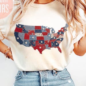 May include: Cream-colored t-shirt featuring a patchwork design of the United States map in red, white, and blue. The shirt includes the text "COMFORT COLORS" in a pink circle. The bottom of the shirt includes the text "LilysunflowerCrafts".