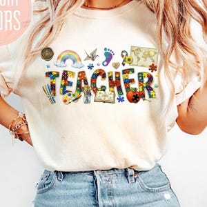 Puede incluir: Camiseta color crema con la palabra "TEACHER" en letras de piezas de rompecabezas de colores. La camiseta presenta un arcoíris, un mapa del mundo y otros gráficos. El texto "COMFORT COLORS" está en un círculo rosa.