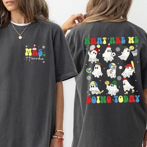 Può includere: Due magliette grigie a tema natalizio. La maglietta anteriore ha il testo "Mrs. Hannah" con uno schema di colori rosso, giallo, blu e verde. La maglietta posteriore ha il testo "What are we doing today?" con un gruppo di fantasmi che praticano sport. I fantasmi indossano cappelli di diversi colori e tengono diversi attrezzi sportivi.