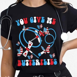 May include: Black t-shirt with the text "YOU GIVE ME BUTTERFLIES" in red, white, and blue. The design includes butterflies and medical equipment in a patriotic color scheme. A stethoscope is draped around the neck.
