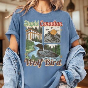 Pode incluir: Uma camiseta azul com um design gráfico com o texto "Stupid Canadian Wolf Bird". O design inclui ilustrações de um edifício, montanhas, um rio e um pássaro. A camiseta tem as mangas enroladas.