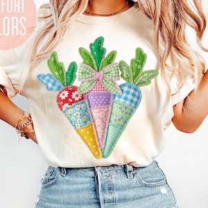 Pode incluir: Camiseta creme com uma aplicação de três cenouras com copas verdes e um laço rosa. As cenouras são feitas de vários tecidos estampados. O texto na camiseta diz "COMFORT COLORS".
