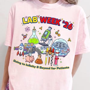 Op de afbeelding: Roze t-shirt met een kleurrijke afbeelding van laboratoriumapparatuur, een raket en een varken. De tekst luidt "LAB WEEK '26" en "Going to Infinity & Beyond for Patients". Een leuk, wetenschappelijk ontwerp.