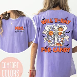Puede incluir: Camiseta morada con el texto "MRS. Hannah" en la parte delantera y "WILL X-RAY FOR CANDY" en la parte trasera. El diseño de la parte trasera presenta un esqueleto con caramelos y fantasmas.