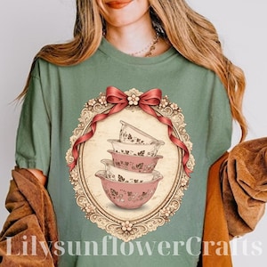 May include: Sage green t-shirt featuring a vintage-style illustration of four pink and white mixing bowls. The bowls are stacked and decorated with floral patterns, set within an ornate oval frame with a red bow. The text "LilysunflowerCrafts" is at the bottom.
