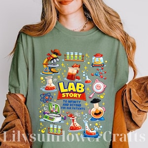 Puede incluir: Camiseta verde salvia con un colorido gráfico de "Lab Story". El diseño presenta equipos de laboratorio, un zorro y el texto "To infinity and beyond for our patients". El gráfico es en tonos de rojo, amarillo, azul y blanco.