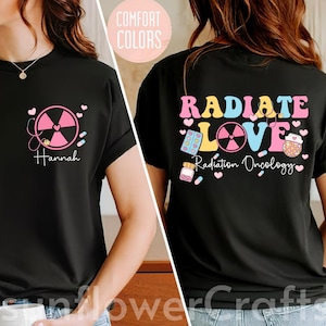 May include: Black t-shirt with a pink and yellow graphic that says "RADIATE LOVE Radiation Oncology". The graphic includes a pink radiation symbol, pills, and a heart.