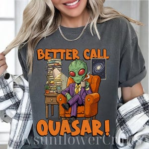 Może przedstawiać: Ciemnoszara koszulka z pomarańczowym napisem "BETTER CALL QUASAR!". Grafika przedstawia kosmitę w fioletowym garniturze, siedzącego w pomarańczowym fotelu, trzymającego kubek. Na koszulce znajduje się również stos książek i obraz galaktyki.