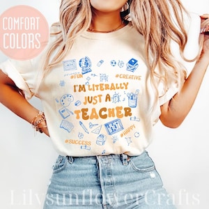 May include: Cream-colored t-shirt with the text "I'M LITERALLY JUST A TEACHER" in orange and blue. The shirt features blue doodles of school supplies and words like "Fun," "Creative," and "Happy."