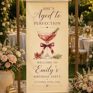 May include: A vertical cream-colored fabric banner hanging from a wooden frame. The text reads: SHE'S Aged to PERFECTION, WELCOME TO Emily's BIRTHDAY PARTY, OCTOBER 20TH, 2026. It features a watercolor illustration of a cocktail glass with a bow.