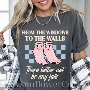 以下が含まれることがあります： 「FROM THE WINDOWS TO THE WALLS」の文字と、顔の描かれたピンク色の靴下の漫画イラストがプリントされた濃いグレーのTシャツ。「There better not be any falls」の文字が靴下の下にあります。