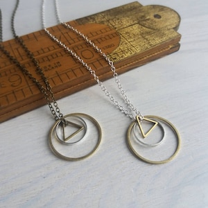 Puede incluir: Dos collares con un diseño geométrico. Un collar tiene una cadena plateada y un círculo plateado con un triángulo plateado en su interior. El otro collar tiene una cadena de bronce y un círculo de bronce con un triángulo de bronce en su interior.