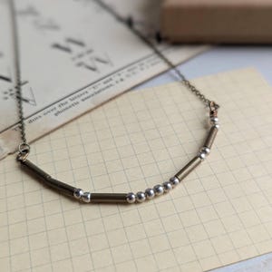 May include: A delicate necklace featuring a curved bar design with silver beads and bronze-colored tubes. The necklace is displayed on a vintage paper background, showcasing its minimalist and elegant style. The chain is a fine, delicate link.