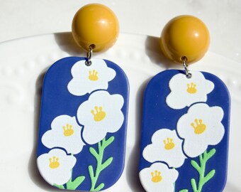 May include: A pair of blue and white floral earrings with yellow centers. The earrings are made of polymer clay and have a gold ball at the top.