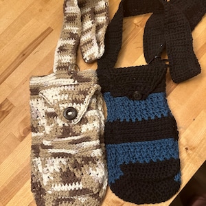 May include: Two handmade crochet bags. One bag is a mix of brown, beige, and white, with a button closure. The other is black with blue stripes and a button closure. Both bags have long straps.