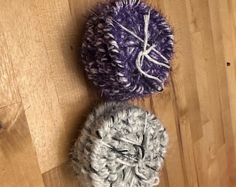 May include: Two handmade yarn balls. One is purple and white, the other is gray and white. Both are round and have white yarn wrapped around them. The yarn balls are sitting on a wooden surface.