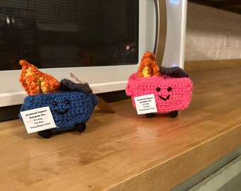May include: Two crocheted stuffed toys shaped like dumpsters with flames on top. The dumpster on the left is blue with a tag that reads "Emotional Support Dumpster Fire It's fine. I'm fine. Everything is fine!" The dumpster on the right is pink with a tag that reads "Emotional Support Dumpster Fire It's fine. I'm fine. Everything is fine!"