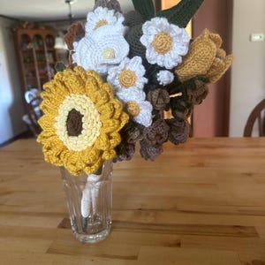 May include: A crochet bouquet in a clear glass vase. The bouquet features a large yellow sunflower, white daisies, and other flowers in shades of yellow, brown, and green. The vase sits on a wooden table.