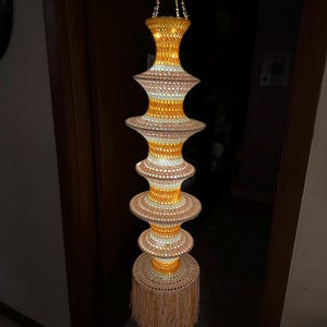 May include: A hanging crochet light fixture featuring a vertical stack of six circular, tiered sections in alternating shades of yellow and cream. The structure is suspended by yellow cords and finished with a long, cream-coloured fringe at the base.