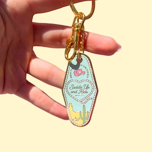 Pode incluir: Um chaveiro dourado com uma etiqueta de chave de motel azul e branca. A etiqueta de chave apresenta um chap&eacute;u de cowboy rosa, um cora&ccedil;&atilde;o com o texto "Saddle Up and Ride" e uma cena do deserto com um cacto e um la&ccedil;o.