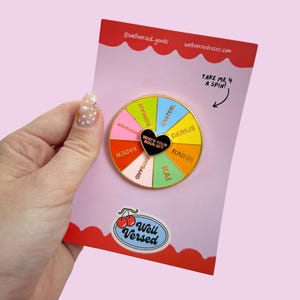 Könnte beinhalten: Ein farbenfroher Emaille-Pin mit einem Glücksrad-Design, das Charakternamen und den Text "Who's Your Book BF?" auf einer rosa Karte zeigt. Die Karte trägt den Text "Take Me 4 A Spin!" und das Well Versed-Logo.