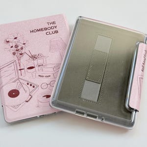 May include: A pink tablet case and cover with a clear plastic frame. The cover has a glittery finish and features a line drawing of a lamp, flowers, and a record player. The text "THE HOMEBODY CLUB" is printed on the cover.