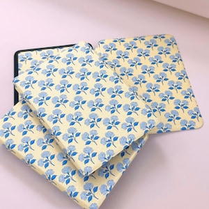 May include: Three book covers with a floral design. The covers display a repeating pattern of blue flowers on a pale yellow backdrop. The covers are stacked, with the top one partially open, revealing a black interior.