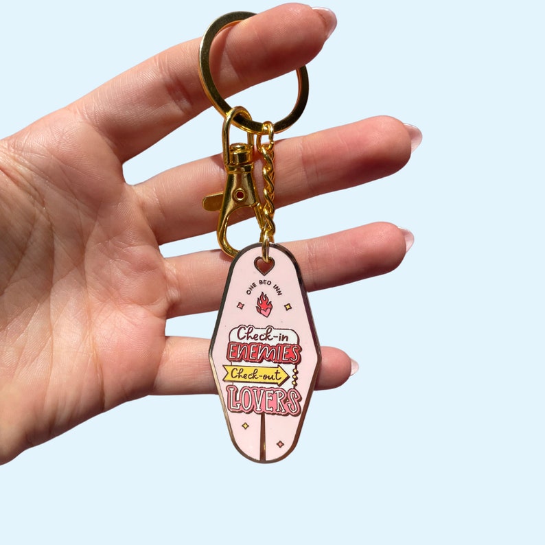 Pode incluir: Chaveiro de motel rosa com detalhes dourados. O chaveiro tem o texto "One Bed Inn" e "Check-in Enemies, Check-out Lovers".