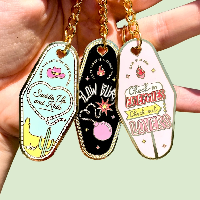 Pode incluir: Tr&ecirc;s chaveiros de motel dourados com designs de esmalte coloridos. O primeiro chaveiro diz "Wear the hat ride the cowboy" com um chap&eacute;u de cowboy rosa e um cacto. O segundo chaveiro diz "Slow burn" com uma bomba rosa e chamas. O terceiro chaveiro diz "One bed inn" com um cora&ccedil;&atilde;o vermelho e diz "Check-in enemies, check-out lovers".