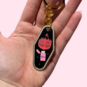 Pode incluir: Um chaveiro de motel preto e dourado com uma chama rosa e o texto "Will they won't they" e "Well Versed".
