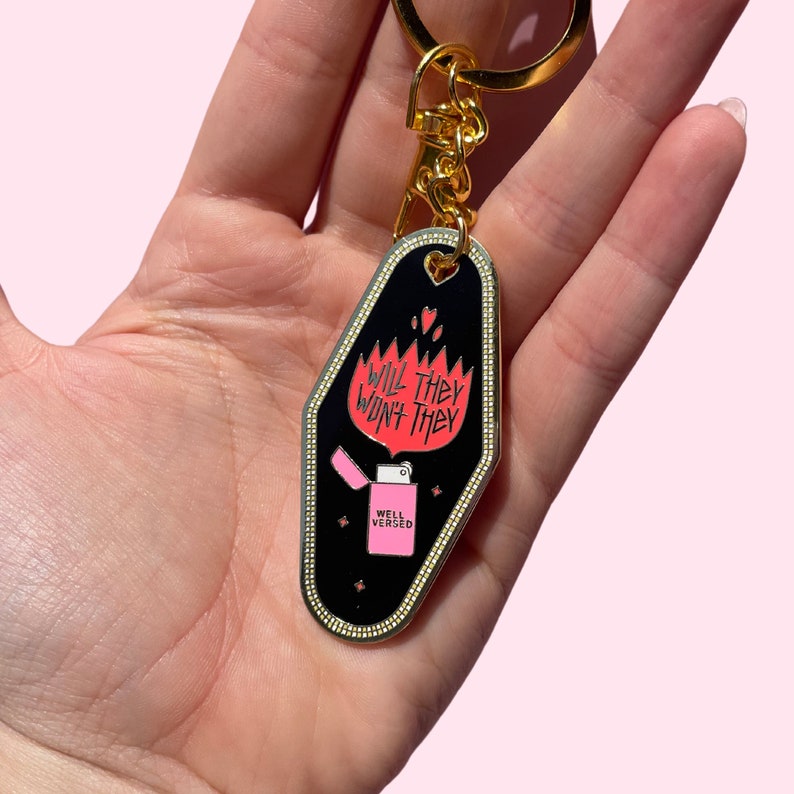 Pode incluir: Um chaveiro de motel preto e dourado com uma chama rosa e o texto "Will they won't they" e "Well Versed".