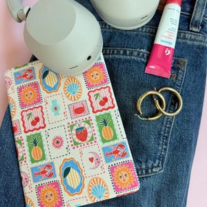 Puede incluir: Una composición sobre unos vaqueros azules: una funda estampada con ilustraciones de frutas y soles, unos auriculares blancos, un tubo de brillo labial rosa y pendientes de aro dorados. El fondo es rosa claro.