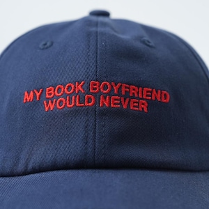 May include: A navy blue baseball cap featuring the embroidered text MY BOOK BOYFRIEND WOULD NEVER in bold red capital letters across the front panels. The cap has a structured crown with a central seam and matching navy blue stitching throughout.