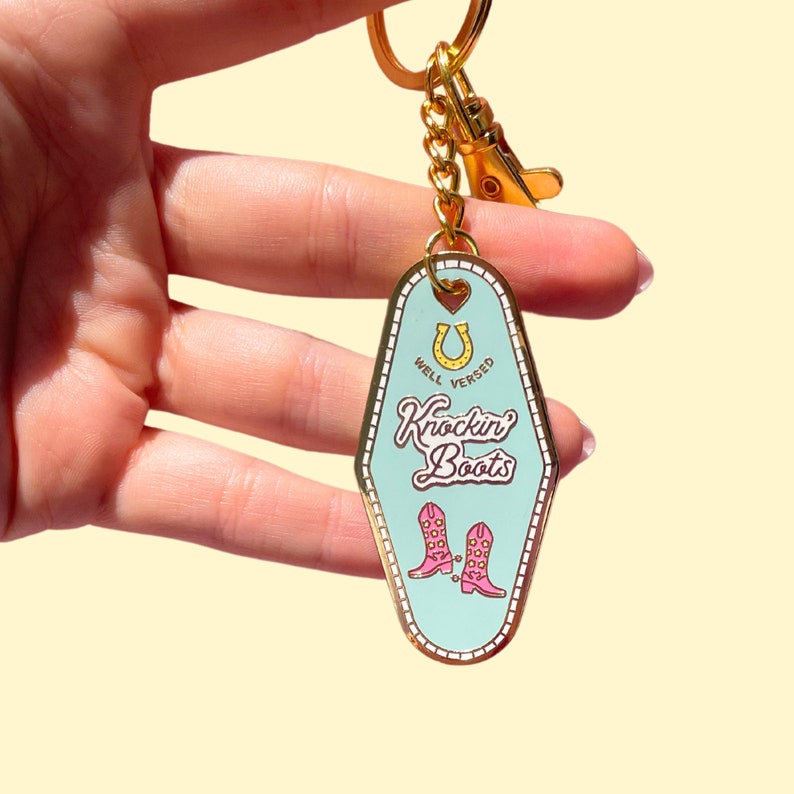 Pode incluir: Um chaveiro dourado com uma etiqueta de chave de motel azul turquesa. A etiqueta da chave tem uma ferradura com as palavras "Well Versed" escritas ao redor. O texto "Knockin' Boots" est&aacute; escrito em uma fonte retr&ocirc;. Duas botas de cowboy rosa com fivelas douradas s&atilde;o representadas abaixo do texto.
