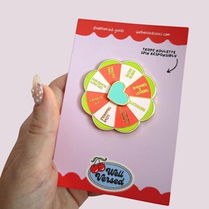 May include: A pin with a wheel design, featuring phrases such as "Grumpy Sunshine" and "Love Triangle." The pin is on a pink card with the words "Well Versed" and "Trope Roulette Spin Responsibly."
